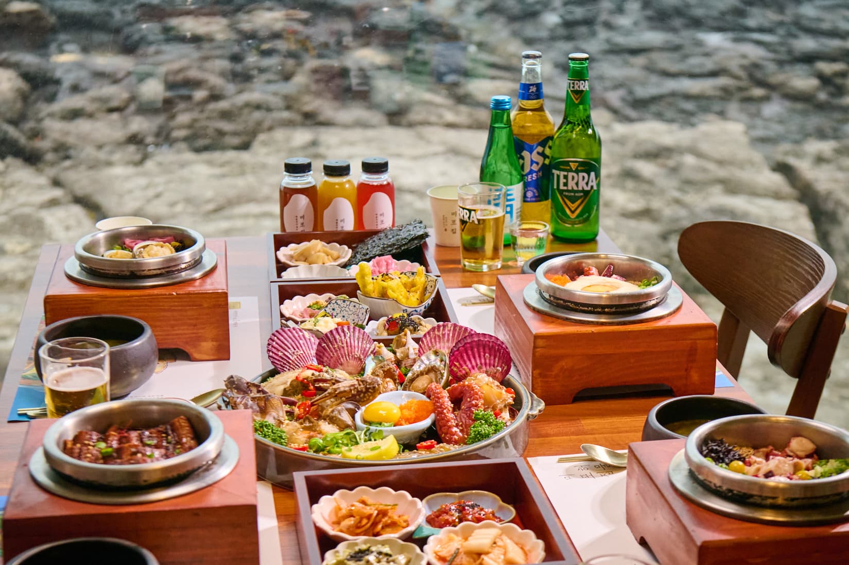 통창 너머 갯바위가 보이는 자리에 해물장 접시와 솥밥 솥, 찬합과 음료가 한 상으로 차려진 모습