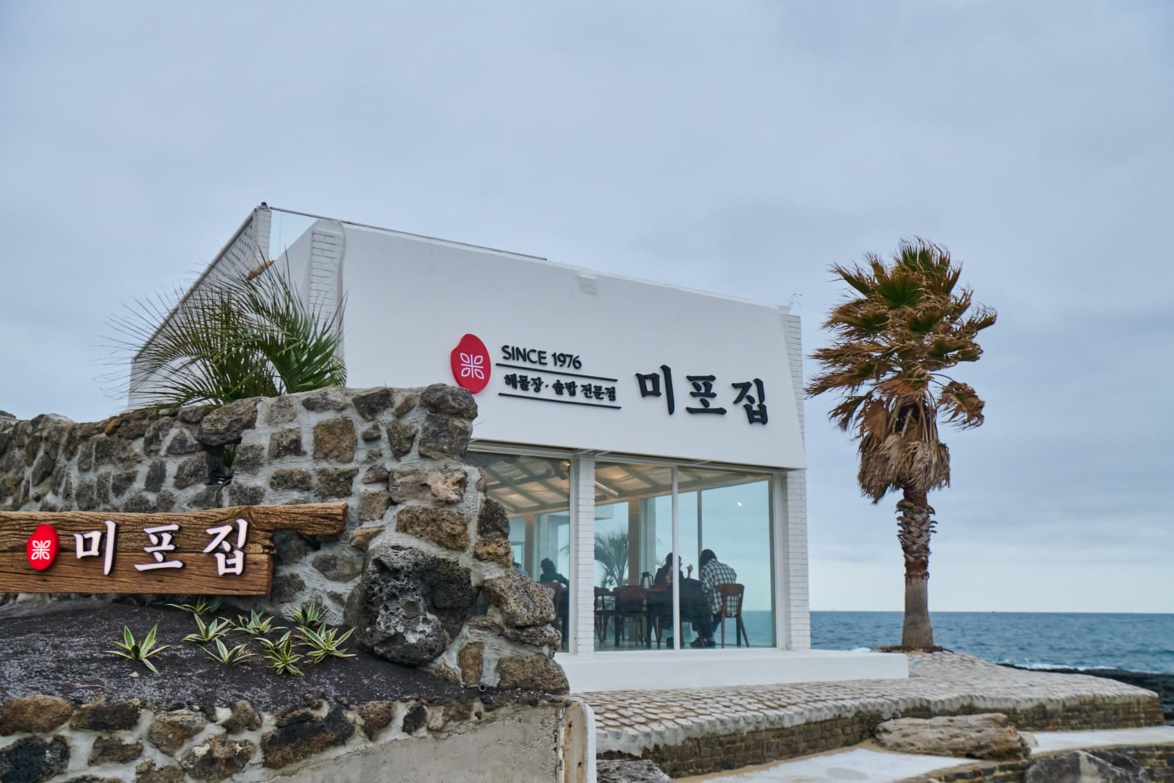 돌담 위 미포집 간판과 야자수, 그 뒤로 이어지는 바다