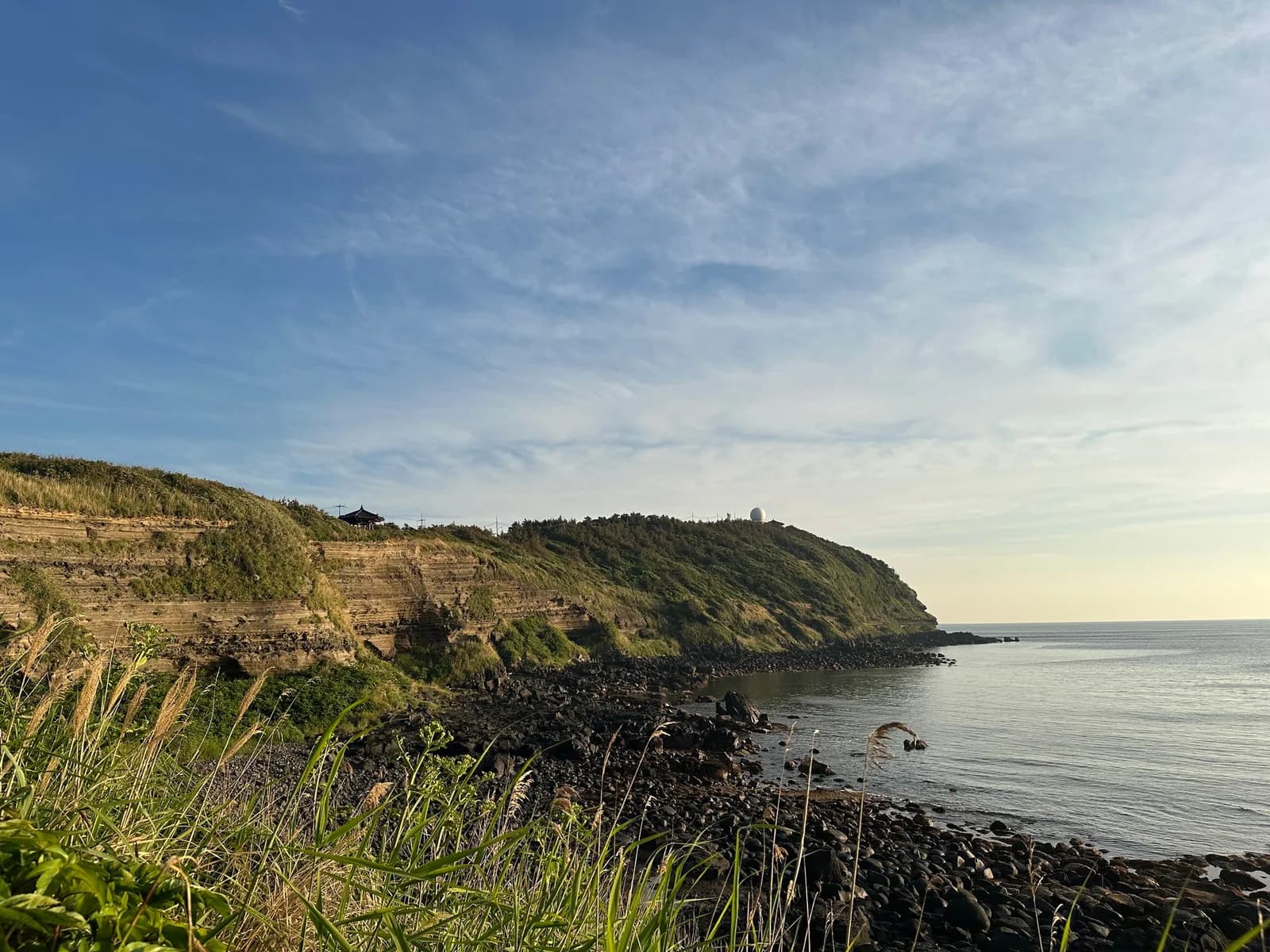 Suwolbong's layered cliff, with a pavilion and a white dome on top, beyond a pebble shore in the evening light
